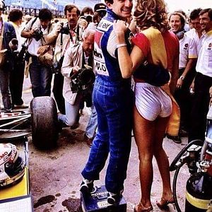 Alain Prost stands on a mechanic's box with an arm around a pit girl in front of a sea of photographers